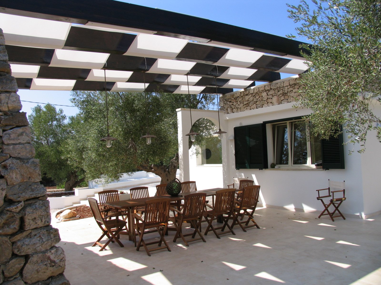 Villa B a Santa Maria di Leuca: portico con pergola in travi scure e tendaggi bianchi, tavolo da pranzo in legno e muro in pietra a secco tra gli ulivi